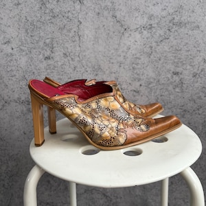 May include: A pair of brown leather mules with a pointed toe and high, square heels. The uppers feature a floral pattern with tan and cream accents. The interior is lined with red leather.