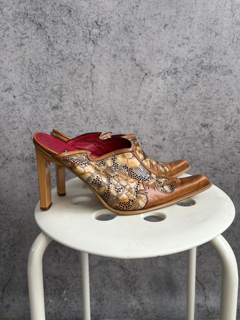 May include: A pair of brown leather mules with a pointed toe and high, square heels. The uppers feature a floral pattern with tan and cream accents. The interior is lined with red leather.