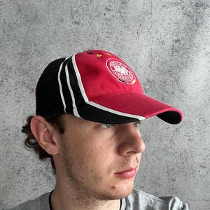 May include: A baseball cap featuring a red front panel with the Deutscher Fussball-Bund logo and two yellow stars, black side panels with three white stripes, and a red brim with a black underside. The cap has a structured crown and eyelets.