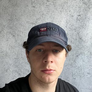 Könnte beinhalten: Marineblaue Diesel Division Baseballkappe mit gebogenem Schirm. Die Kappe hat einen gestickten Text und ein Logo auf der Vorderseite. Der Hut ist aus einem strukturierten Stoff gefertigt.