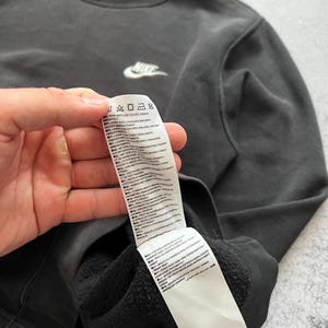 May include: A dark gray Nike sweatshirt with a white Nike logo on the chest. The sweatshirt has a crew neck and a tag with care instructions. The fabric appears to be a soft, comfortable material.