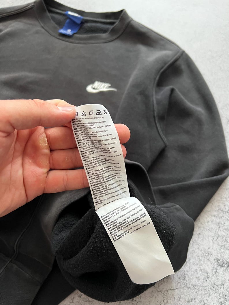 May include: A dark gray Nike sweatshirt with a white Nike logo on the chest. The sweatshirt has a crew neck and a tag with care instructions. The fabric appears to be a soft, comfortable material.