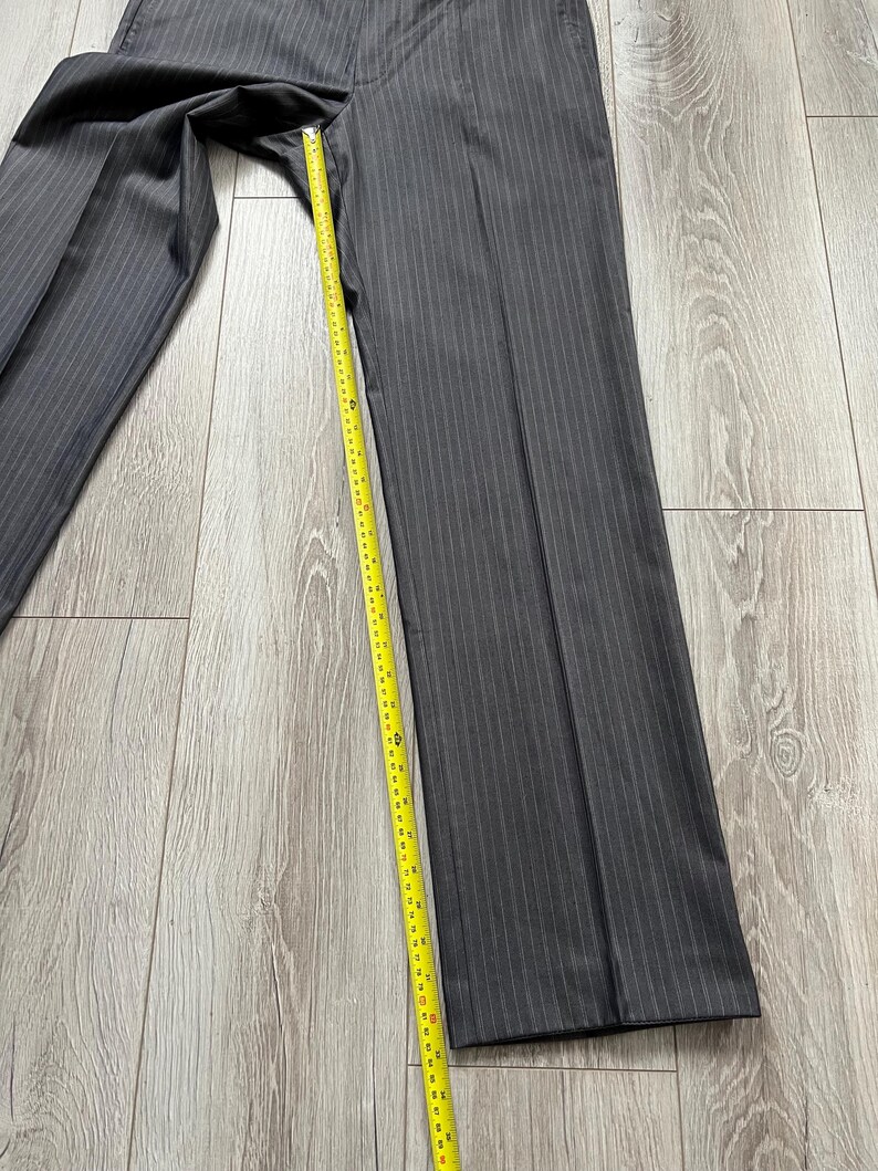 May include: Dark gray trousers with a subtle vertical pinstripe pattern. A yellow measuring tape is placed along the inner seam, showing a length measurement extending from the crotch to the hem, reaching approximately 32 inches in total.