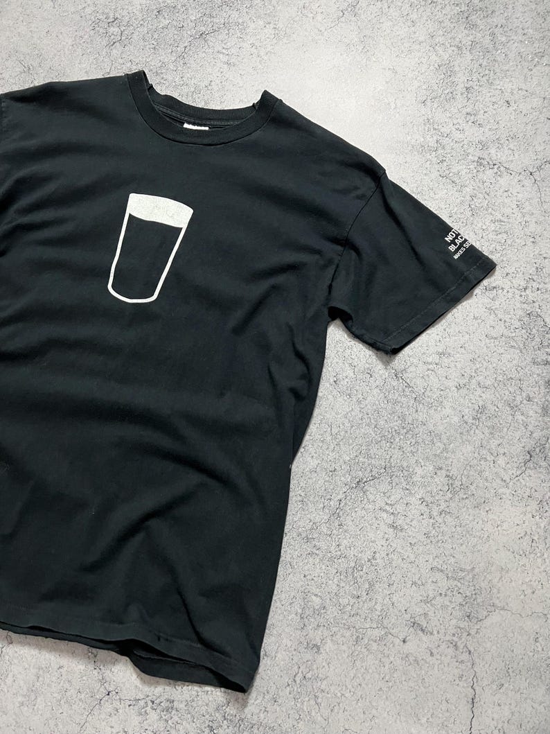 May include: Black t-shirt featuring a white outline of a pint glass. The shirt has a crew neck and short sleeves. The text "NOT BLACK MAKES SENSE" is printed on the sleeve.