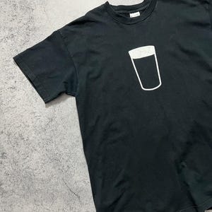 May include: Black t-shirt featuring a white outline of a pint glass. The graphic is centered on the chest. The shirt has short sleeves and a crew neck. The fabric appears to be cotton.