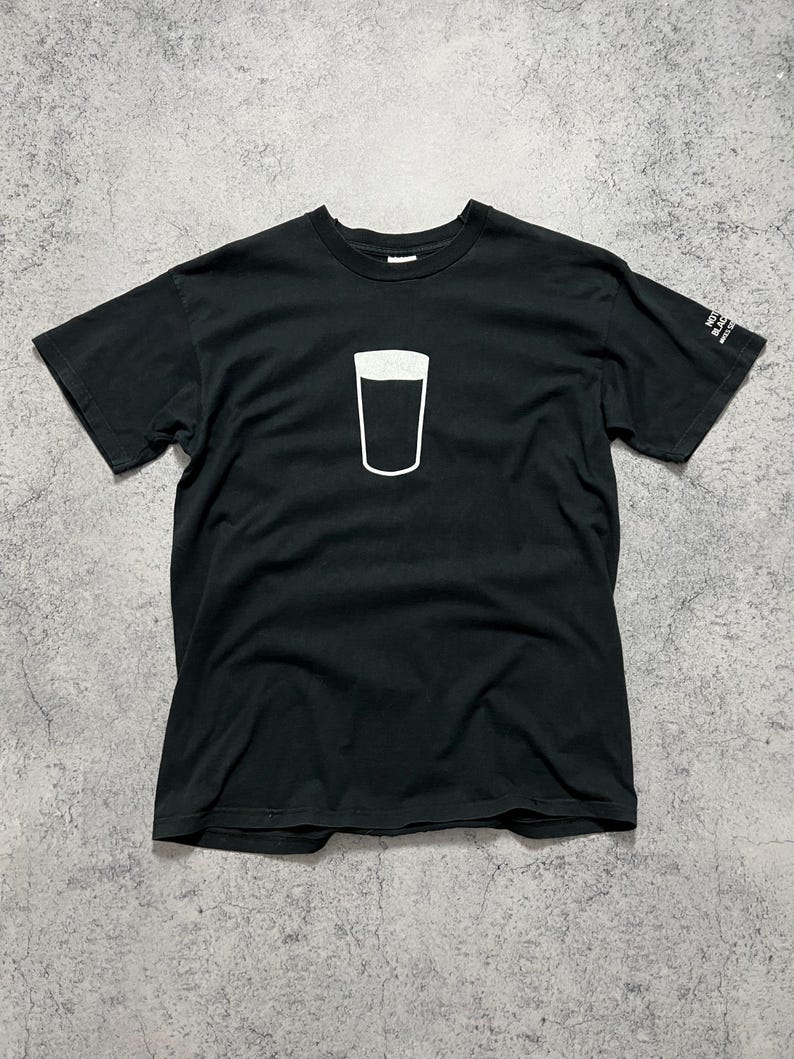 May include: Black t-shirt featuring a white outline of a pint glass. The shirt has short sleeves and a crew neck. The text on the sleeve is blurry.