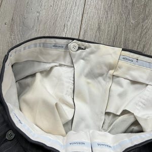 May include: Dark gray pinstriped trousers featuring a white interior waistband lining with the repeated text "VERSACE" in blue. The trousers include a single white button closure at the center front and show visible yellowing on the inner fabric.