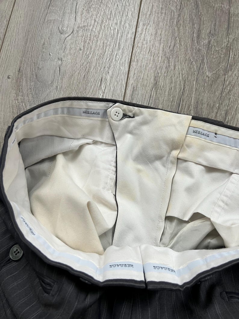 May include: Dark gray pinstriped trousers featuring a white interior waistband lining with the repeated text "VERSACE" in blue. The trousers include a single white button closure at the center front and show visible yellowing on the inner fabric.