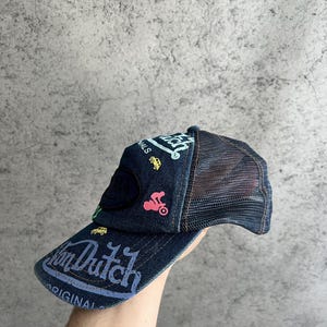 May include: A dark blue denim trucker hat featuring a mesh back and a curved brim. The front panel displays the text Von Dutch and Original in light blue, accented by small embroidered graphics of a yellow car and a pink motorcycle.