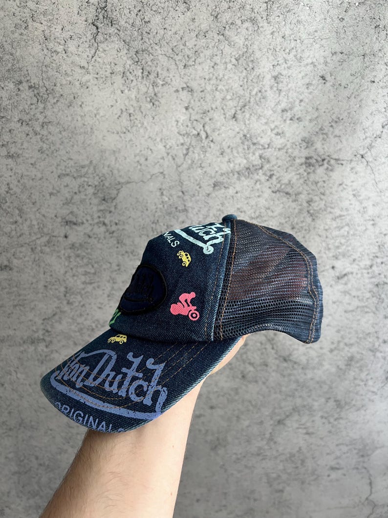 May include: A dark blue denim trucker hat featuring a mesh back and a curved brim. The front panel displays the text Von Dutch and Original in light blue, accented by small embroidered graphics of a yellow car and a pink motorcycle.