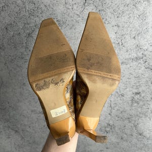 May include: A pair of tan high heels with pointed toes and a snakeskin pattern. The heels have a glossy finish and a small, square heel. The soles are worn and show signs of use.