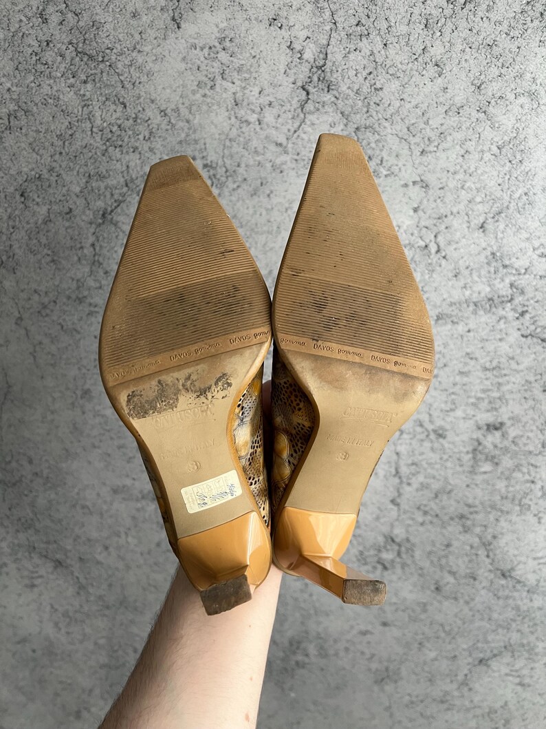 May include: A pair of tan high heels with pointed toes and a snakeskin pattern. The heels have a glossy finish and a small, square heel. The soles are worn and show signs of use.