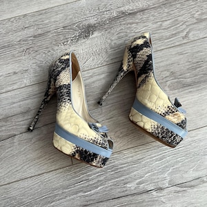 May include: A pair of high-heeled platform pumps featuring a snakeskin pattern in cream, black, and gray. The shoes have a light blue trim around the platform and a small bow detail at the peep-toe front. The interior lining is beige.