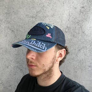 May include: A blue denim trucker hat featuring a dark blue mesh back and a curved brim. The front panel displays a dark oval patch with the text Von Dutch, alongside various colorful embroidered graphics including a car and a motorcycle.