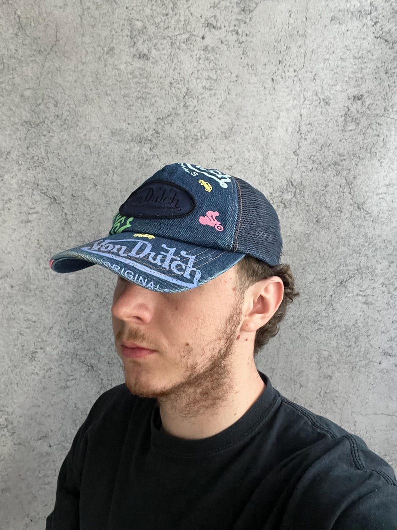 May include: A blue denim trucker hat featuring a dark blue mesh back and a curved brim. The front panel displays a dark oval patch with the text Von Dutch, alongside various colorful embroidered graphics including a car and a motorcycle.