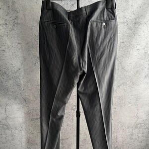 May include: A pair of dark gray trousers featuring a subtle vertical pinstripe pattern, a waistband with belt loops, and two rear pockets with button closures. The trousers are displayed on a black plastic hanger and a black metal stand.