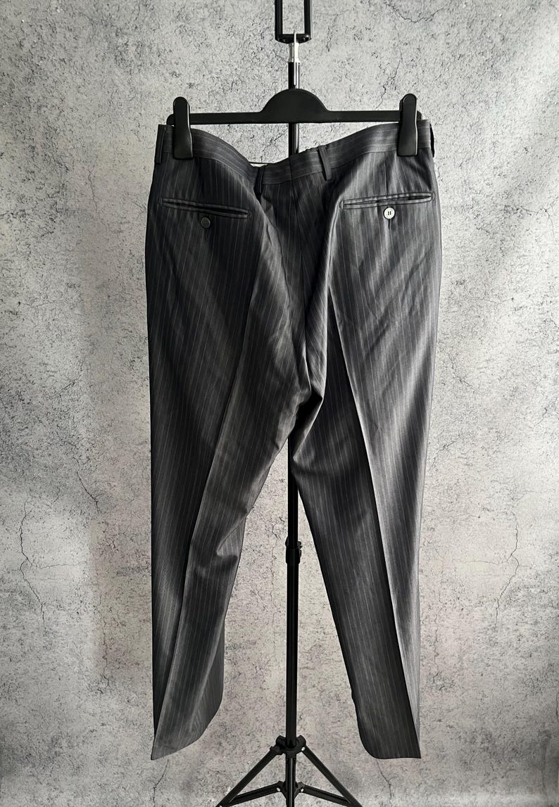 May include: A pair of dark gray trousers featuring a subtle vertical pinstripe pattern, a waistband with belt loops, and two rear pockets with button closures. The trousers are displayed on a black plastic hanger and a black metal stand.
