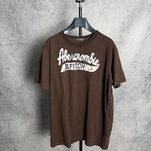 May include: A brown Abercrombie & Fitch t-shirt with a white logo. The short-sleeved shirt is hanging on a black hanger against a grey background. The logo is in a script font with a white outline.