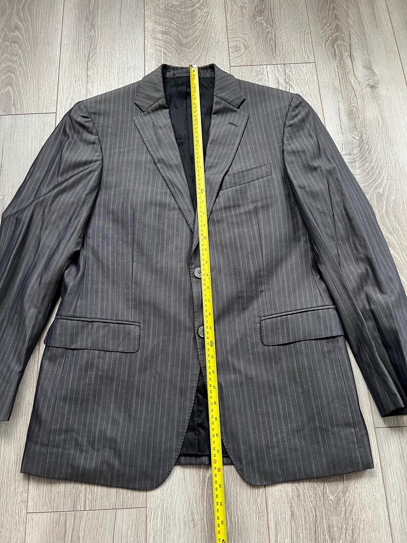 May include: A charcoal gray pinstriped blazer with a notched lapel, two front buttons, and two flap pockets. A yellow measuring tape is placed vertically over the center, showing a length of approximately 30 inches from collar to hem.