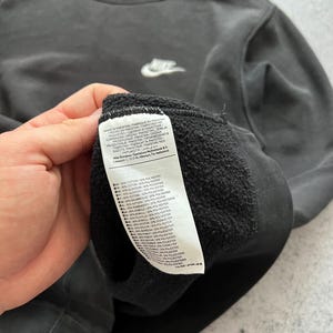 May include: A dark gray Nike sweatshirt with a small white Nike logo on the chest. The sweatshirt has a crew neck and a small blue tag on the back of the neck. A white tag with text is visible.
