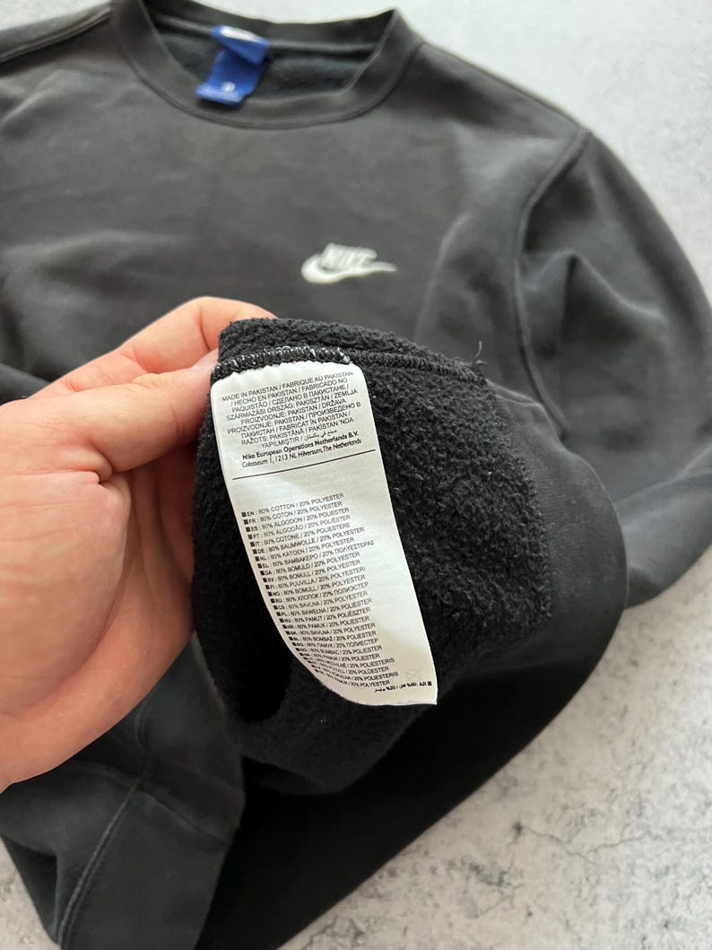 May include: A dark gray Nike sweatshirt with a small white Nike logo on the chest. The sweatshirt has a crew neck and a small blue tag on the back of the neck. A white tag with text is visible.