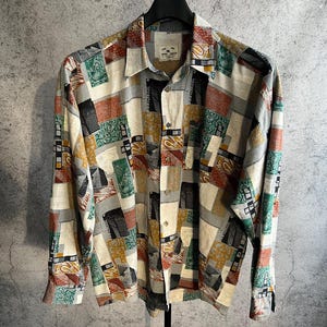 May include: A patterned long-sleeved shirt with a button-down front. The shirt displays a patchwork design with various colors, including red, green, yellow, and black. It hangs on a black hanger against a grey backdrop.