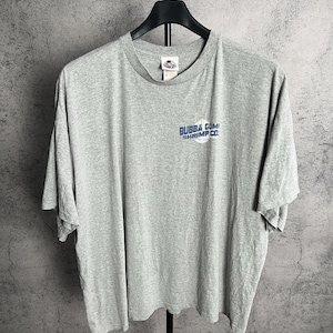 Könnte beinhalten: Ein hellgraues T-Shirt mit dem blauen Schriftzug "Bubba Gump Shrimp Co.". Das Shirt hängt an einem schwarzen Kleiderbügel vor einem grauen Hintergrund. Es hat kurze Ärmel und einen Rundhalsausschnitt.