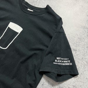 May include: Black t-shirt featuring a white outline of a pint glass. The sleeve displays the text "NOT EVERYTHING IN BLACK AND WHITE MAKES SENSE GUINNESS" in white. A casual, everyday wear tee.
