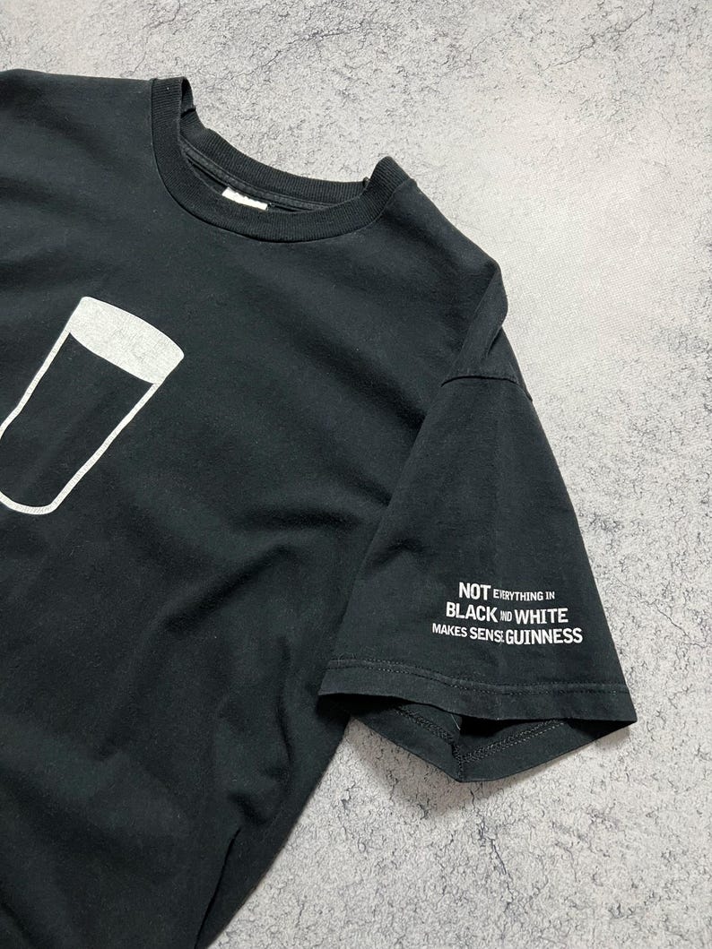 May include: Black t-shirt featuring a white outline of a pint glass. The sleeve displays the text "NOT EVERYTHING IN BLACK AND WHITE MAKES SENSE GUINNESS" in white. A casual, everyday wear tee.