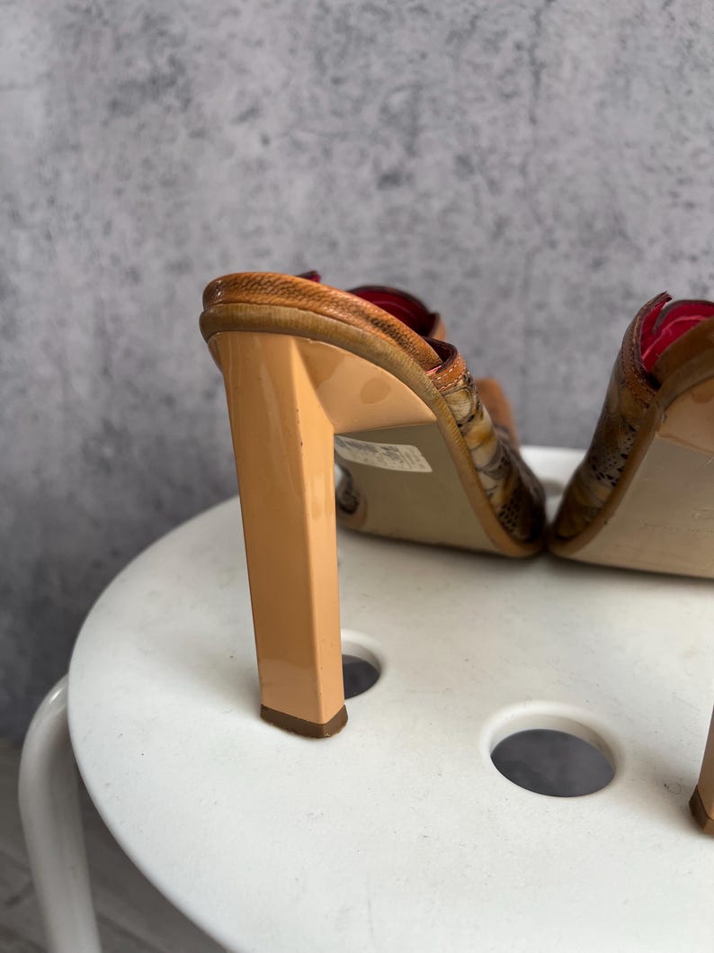 May include: A pair of high-heeled sandals with a snakeskin pattern. The heels are a light tan color, and the soles are a darker brown. The sandals have a red interior.