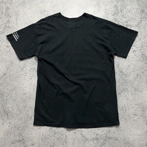 May include: Black t-shirt with short sleeves. The text "WHITE GUINNESS" is printed in white on the left sleeve. The shirt is made of a soft, dark fabric and is laid flat on a textured surface.