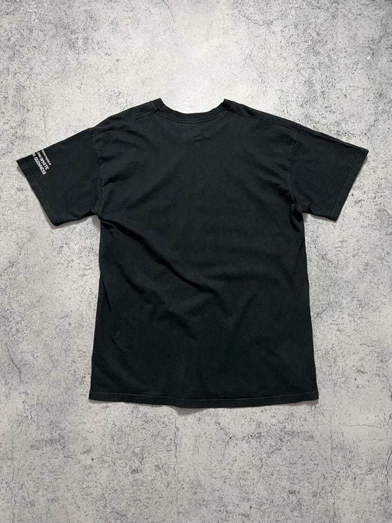 May include: Black t-shirt with short sleeves. The text "WHITE GUINNESS" is printed in white on the left sleeve. The shirt is made of a soft, dark fabric and is laid flat on a textured surface.