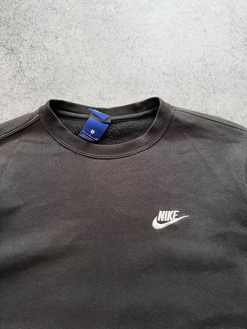 May include: Black crew neck sweatshirt with the Nike logo embroidered in white on the chest. The sweatshirt has a tag attached near the collar. The fabric appears to be a soft, comfortable material.