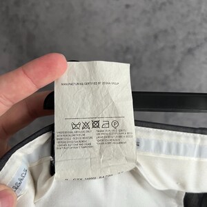 May include: A white fabric care label attached to a dark garment, featuring the text Manufacturing Certified by Zegna Group. The label includes laundry symbols and instructions for professional dry cleaning and warm ironing at 110 C or 230 F.