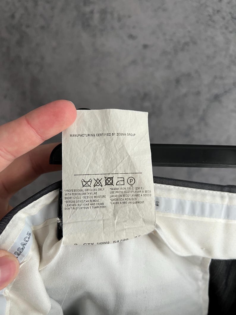 May include: A white fabric care label attached to a dark garment, featuring the text Manufacturing Certified by Zegna Group. The label includes laundry symbols and instructions for professional dry cleaning and warm ironing at 110 C or 230 F.
