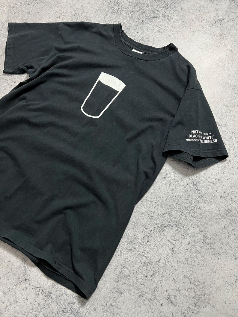 May include: Black t-shirt featuring a white outline of a pint glass with a dark fill. The sleeve has the text "NOT EVERYTHING IN BLACK & WHITE MAKES SENSE GUINNESS".