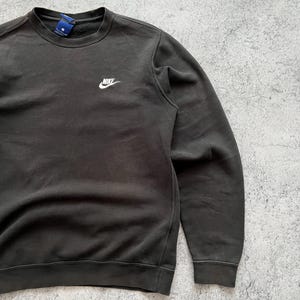 May include: Dark gray crewneck sweatshirt with the Nike logo embroidered in white on the upper left chest. The sweatshirt has a ribbed collar, cuffs, and hem. The fabric appears to be a soft, worn material.