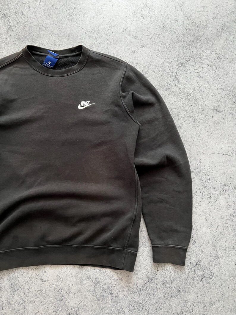 May include: Dark gray crewneck sweatshirt with the Nike logo embroidered in white on the upper left chest. The sweatshirt has a ribbed collar, cuffs, and hem. The fabric appears to be a soft, worn material.