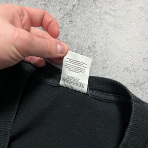 May include: Black t-shirt with a white tag attached. The tag has care instructions and manufacturing information printed on it. The shirt is made of a soft, dark fabric.