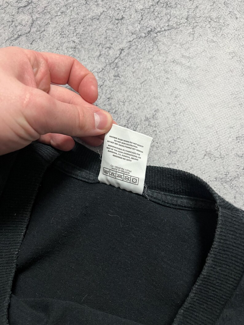 May include: Black t-shirt with a white tag attached. The tag has care instructions and manufacturing information printed on it. The shirt is made of a soft, dark fabric.