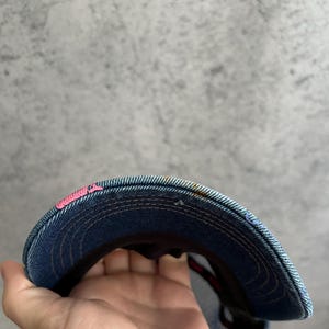 May include: A blue denim baseball cap featuring a curved brim with visible orange stitching and a small patch of bright pink embroidery on the left side. The underside of the brim shows a dark fabric lining and a mesh back panel construction.