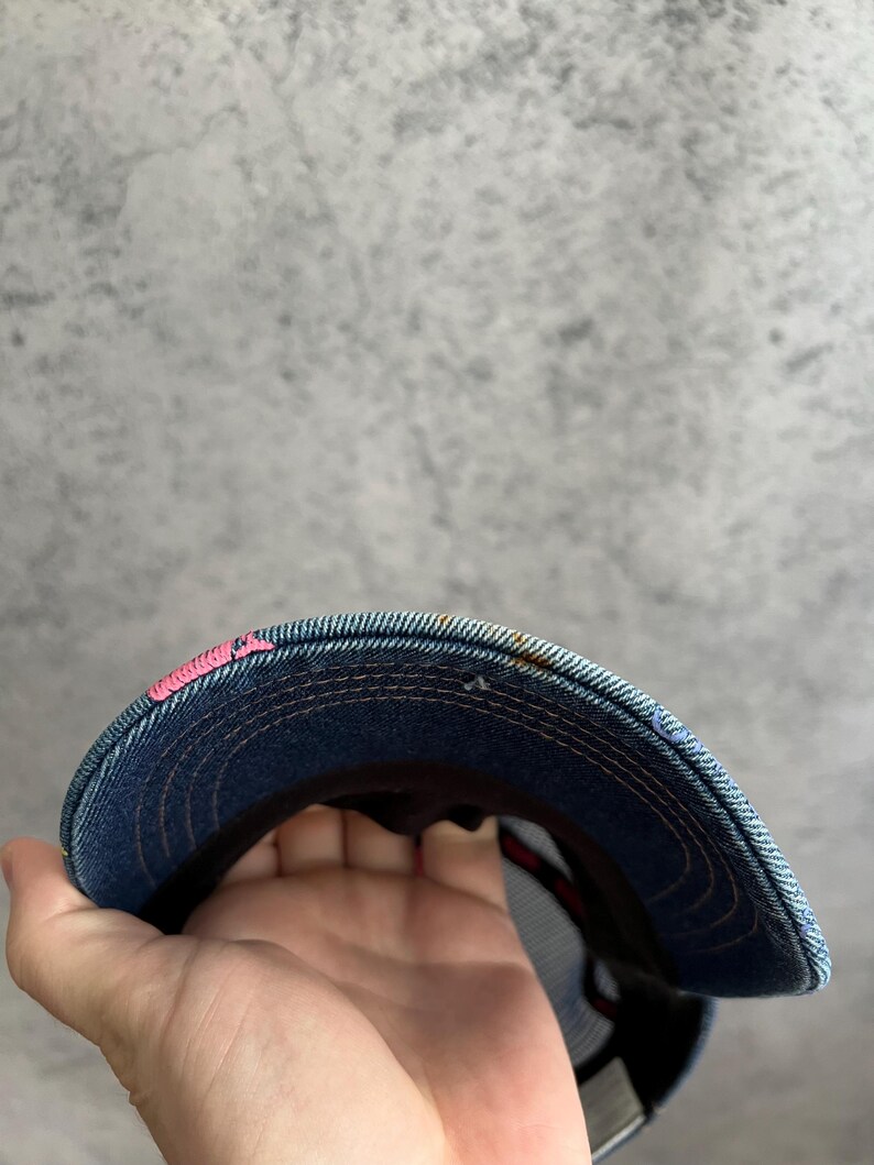 May include: A blue denim baseball cap featuring a curved brim with visible orange stitching and a small patch of bright pink embroidery on the left side. The underside of the brim shows a dark fabric lining and a mesh back panel construction.