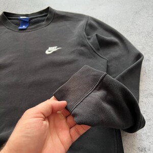 May include: A dark gray Nike crewneck sweatshirt. The sweatshirt features a small white Nike logo on the chest. The cuffs and neckline are ribbed. The fabric appears slightly faded.