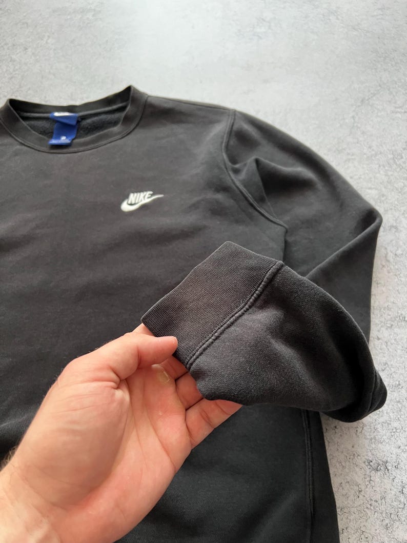 May include: A dark gray Nike crewneck sweatshirt. The sweatshirt features a small white Nike logo on the chest. The cuffs and neckline are ribbed. The fabric appears slightly faded.