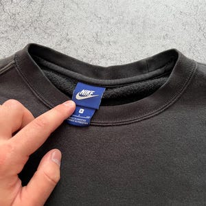 May include: Black crew neck sweatshirt with the Nike logo embroidered in white on the chest and a blue tag at the collar. The tag has the Nike logo and size information. The sweatshirt is a size small.