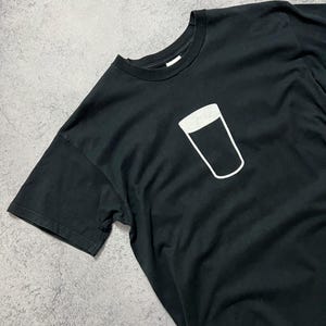 May include: Black t-shirt featuring a white outline of a pint glass. The graphic is centered on the chest. The shirt has short sleeves and a crew neck. The fabric appears to be cotton or a cotton blend.