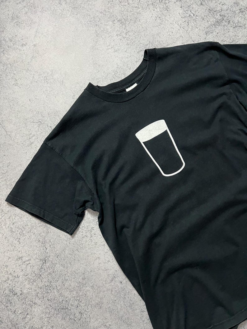 May include: Black t-shirt featuring a white outline of a pint glass. The graphic is centered on the chest. The shirt has short sleeves and a crew neck. The fabric appears to be cotton or a cotton blend.