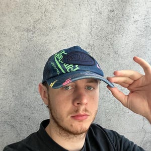May include: A blue denim baseball cap featuring a dark blue patch with the text Von Dutch, accented by bright green and pink graphic lettering and symbols across the front panels and the brim, with a structured crown and a curved visor.