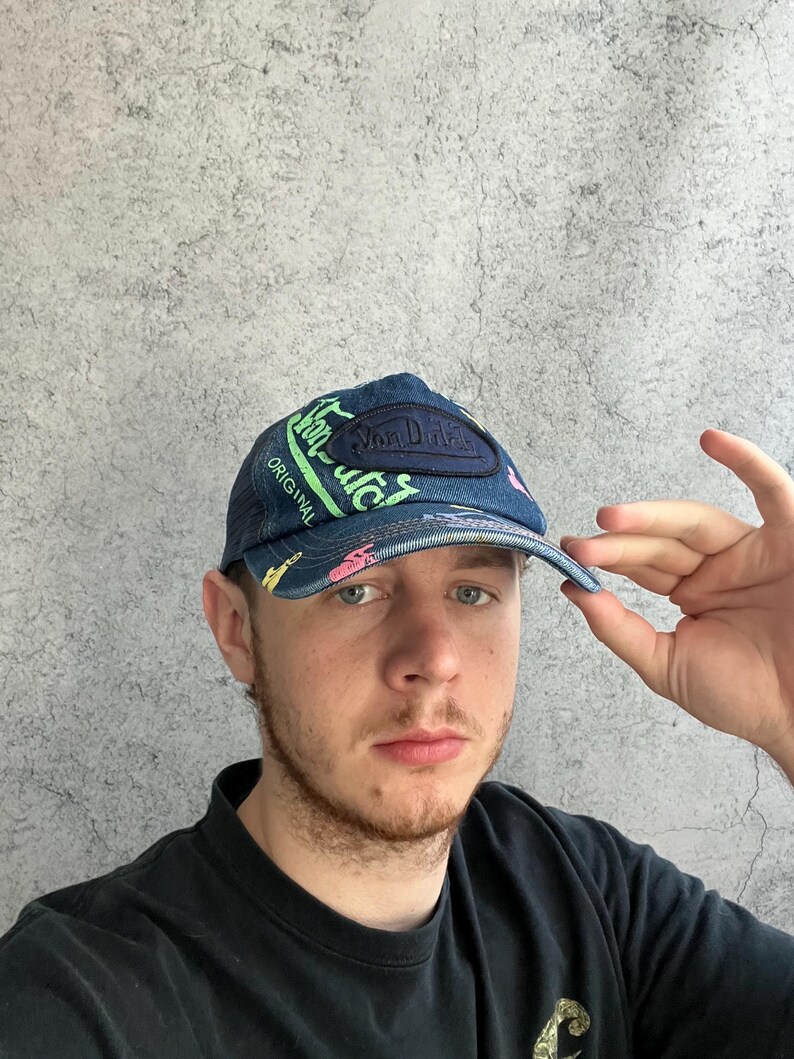 May include: A blue denim baseball cap featuring a dark blue patch with the text Von Dutch, accented by bright green and pink graphic lettering and symbols across the front panels and the brim, with a structured crown and a curved visor.
