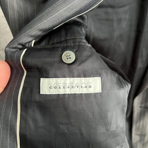 May include: A dark gray pinstriped blazer with a black interior lining featuring a repeating V logo pattern. A white rectangular label reads VERSACE COLLECTION in black text, positioned below a circular gray button with embossed branding.